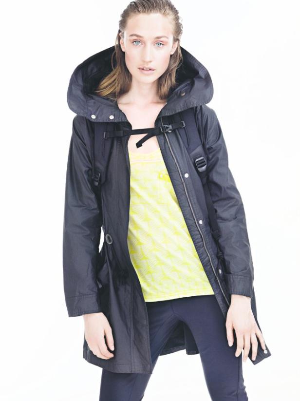 Eine Frau mit Kapuzenjacke und gelbem Oberteil posiert vor einem weißen Hintergrund.