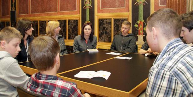 Eine Gruppe von Jugendlichen und Erwachsenen sitzt an einem Tisch in einem formellen Raum.