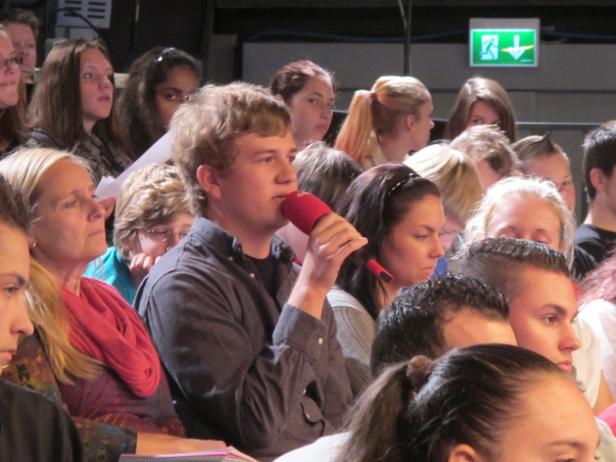 Ein junger Mann spricht in ein rotes Mikrofon vor einem Publikum.