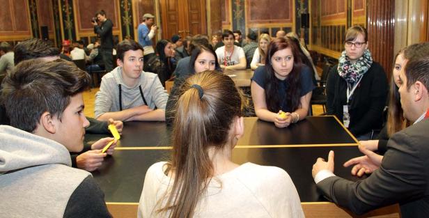 Jugendliche sitzen an Tischen in einem Raum und scheinen an einer Diskussion oder einem Wettbewerb teilzunehmen.