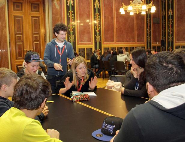 Eine Frau erklärt einer Gruppe von Menschen etwas an einem Tisch in einem verzierten Raum.