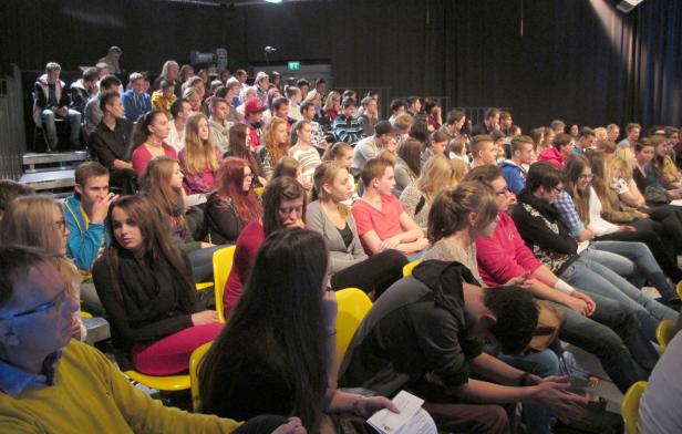 Ein Publikum sitzt auf gelben Stühlen in einem dunklen Raum.
