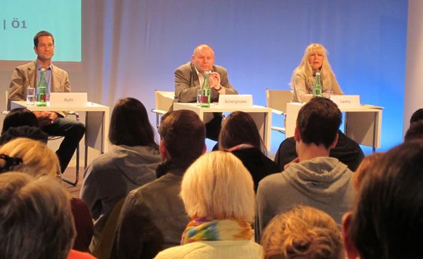 Drei Personen sitzen bei einer Podiumsdiskussion vor Publikum auf der Bühne.