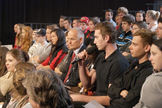 Ein Mann spricht bei einer Veranstaltung in ein Mikrofon, umgeben von Zuhörern.