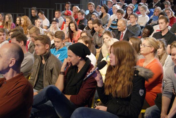 Ein Publikum sitzt bei einer Veranstaltung und hört einer Frau mit Mikrofon zu.