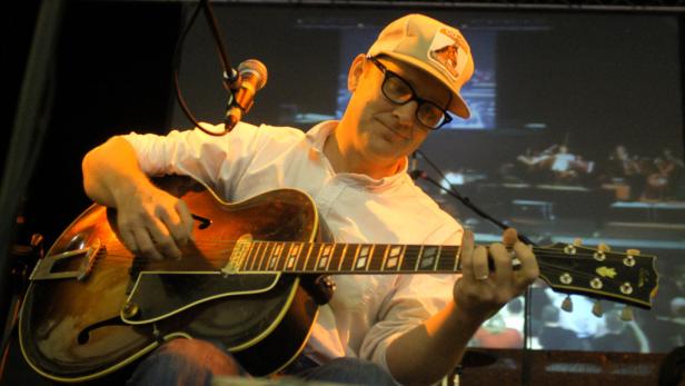 Ein Mann mit Brille und Kappe spielt eine halbakustische Gitarre vor einer Leinwand.