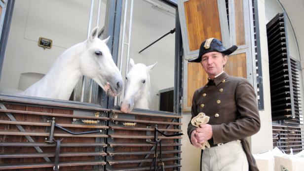 Ein Mann in Uniform steht vor einem Stall mit zwei weißen Pferden.