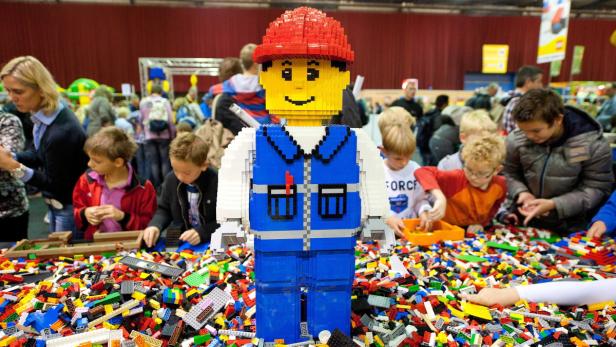Ein großer LEGO-Mann steht inmitten einer Menge von LEGO-Steinen, umgeben von spielenden Kindern.