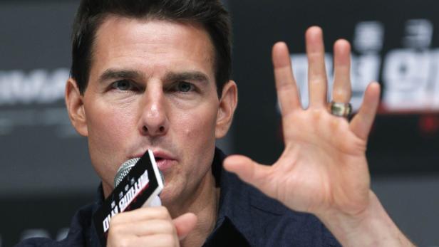 Tom Cruise spricht in ein Mikrofon und hebt seine Hand.