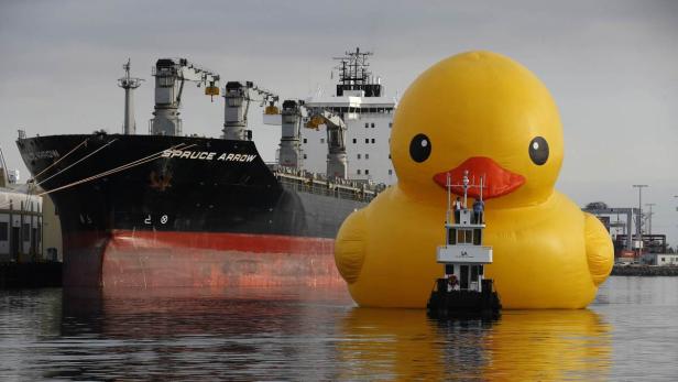 Ein riesige, gelbe Gummiente schwimmt neben einem Frachtschiff im Hafen.