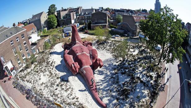 Ein riesiger, liegender Dinosaurier aus Holz in einer städtischen Umgebung.