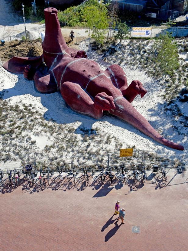 Eine große, rote Dinosaurier-Skulptur liegt auf dem Sand neben einem Fahrradständer.