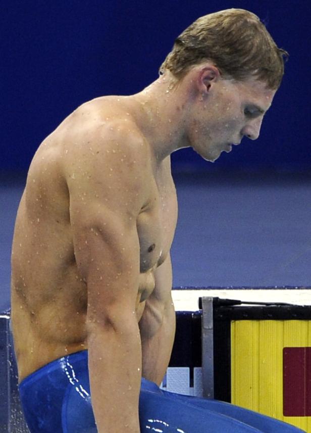 Ein nasser Schwimmer in blauer Badehose steht auf einem Startblock und blickt nach unten.