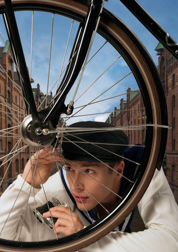 Ein Mann mit Mütze blickt durch das Rad eines Fahrrads mit einem Fernglas in der Hand.