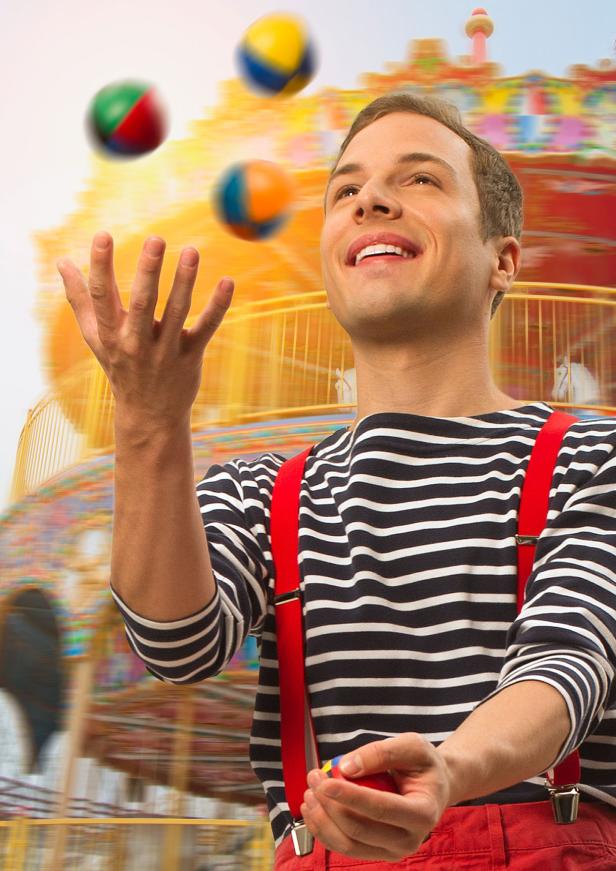 Ein Mann im gestreiften Hemd und Hosenträgern jongliert mit bunten Bällen vor einem Karussell.