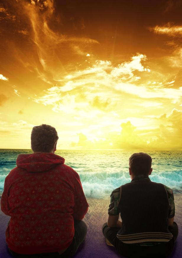 Zwei Männer sitzen am Strand und schauen auf das Meer bei Sonnenuntergang.