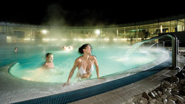 Eine Frau steht in einem beleuchteten Thermalbad, während ein Mann im Hintergrund schwimmt.