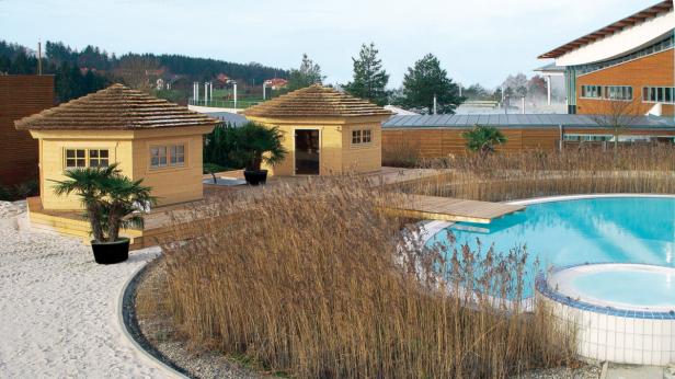 Zwei kleine Holzhäuser mit Strohdach stehen neben einem Pool in einer Wellnessanlage.