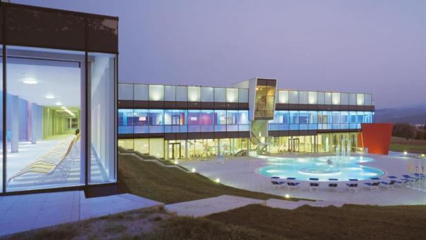 Das moderne Gebäude eines Thermalbads mit Außenpool und Liegestühlen bei Dämmerung.
