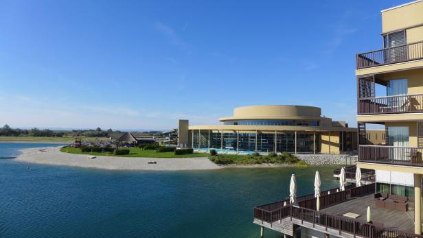 Ein modernes Hotelresort mit einem See und einem Strandbereich an einem sonnigen Tag.