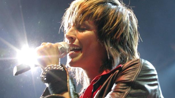 Eine Sängerin mit braunen Haaren singt in ein Mikrofon auf einer Bühne.