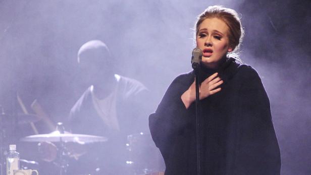 Adele singt in einem dunklen Kleid auf der Bühne, während ein Schlagzeuger im Hintergrund zu sehen ist.