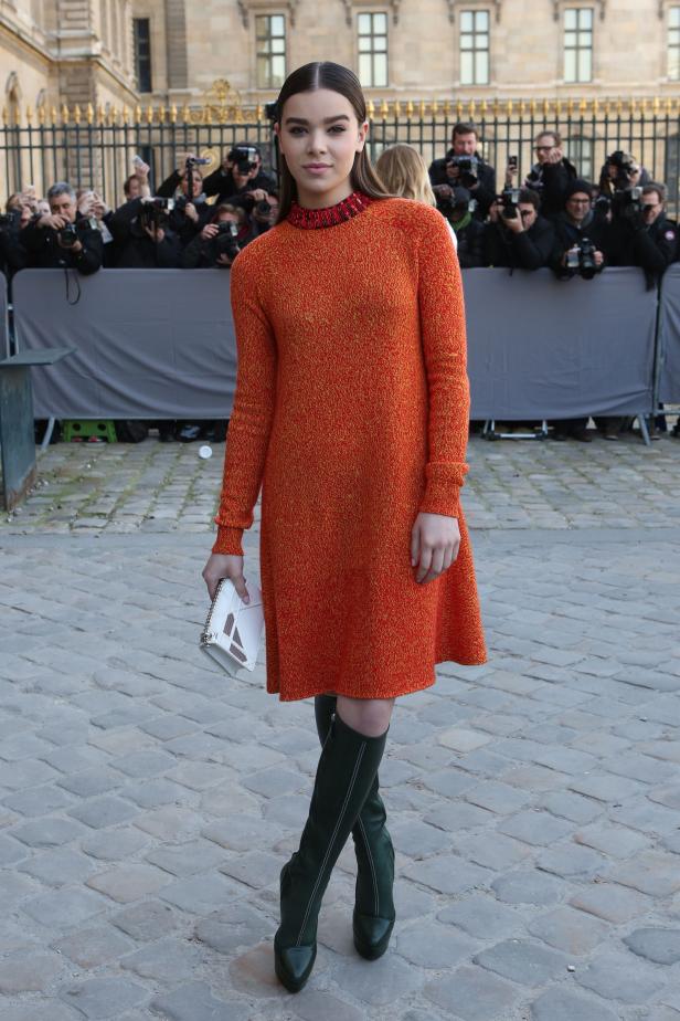 Hailee Steinfeld trägt ein orangefarbenes Kleid und grüne Stiefel vor einer Gruppe von Fotografen.