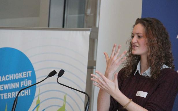 Eine Frau mit lockigem Haar gestikuliert vor einem Mikrofon bei einer Präsentation.