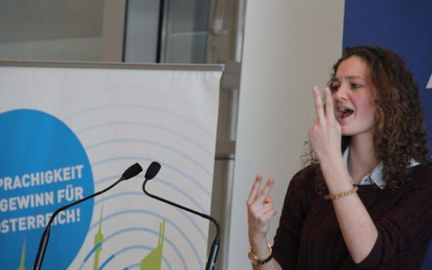 Eine junge Frau gestikuliert vor einem Mikrofon und einem Banner mit der Aufschrift „Sprachigkeit Gewinn für Österreich!“.