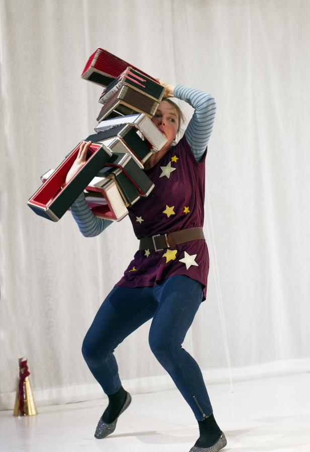 Eine Frau in einem Sternenhemd balanciert einen Stapel Akkordeons auf ihrem Kopf.