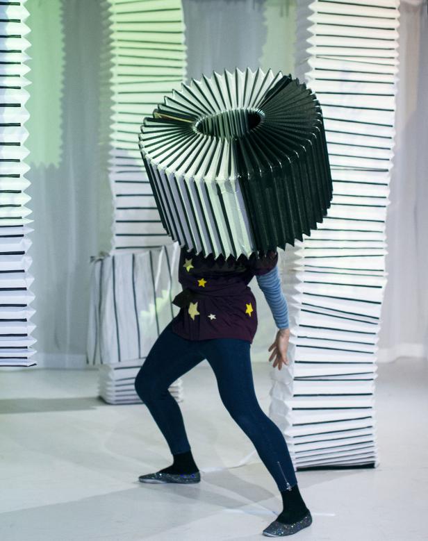 Eine Person mit einem großen, runden, ziehharmonikaartigen Kopfschmuck steht auf einer Bühne.