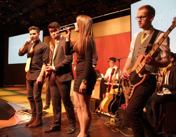 Eine Band tritt auf einer Bühne mit Sängerinnen, Sängern und einem Bassisten auf.