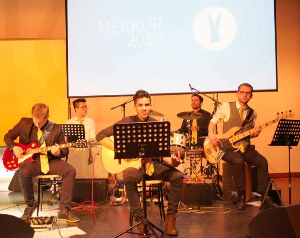 Eine Band spielt auf einer Bühne vor einem Bildschirm mit der Aufschrift „Merkur 2016“.