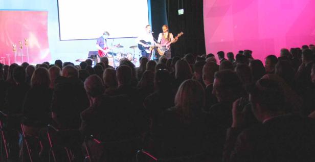 Eine Band spielt vor einem Publikum auf einer Bühne mit pinkfarbener Beleuchtung.