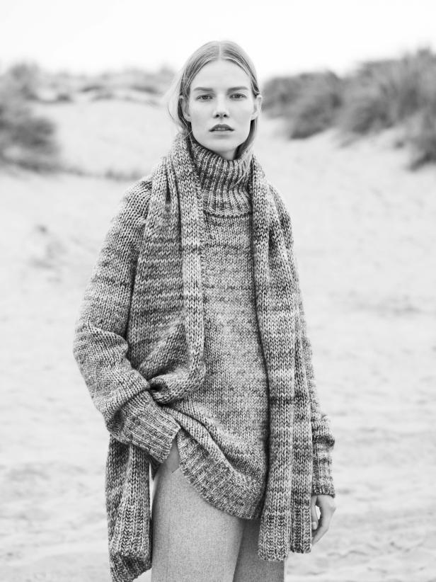 Eine Frau trägt einen übergroßen Strickpullover und einen Schal am Strand.