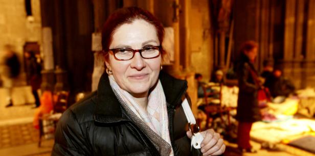 Eine Frau mit Brille und Winterjacke steht in einem Raum mit mehreren Personen.