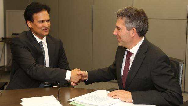 Zwei Männer in Anzügen schütteln sich die Hände über einem Tisch mit Dokumenten.