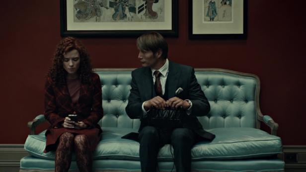 Mads Mikkelsen und eine Frau sitzen auf einem Sofa; sie schaut auf ihr Handy.