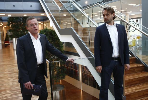 Zwei Männer in Anzügen stehen in einem modernen Gebäude mit Treppenhaus.