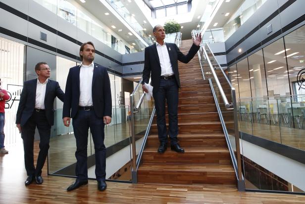 Drei Männer in Anzügen stehen in einem modernen Bürogebäude auf einer Treppe.