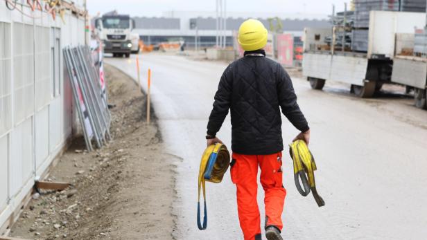 Ein Bauarbeiter mit gelbem Helm und Gurten geht auf einer Baustelle entlang.