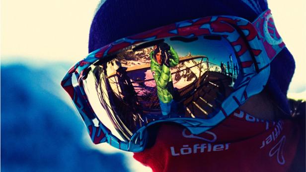 Die Skibrille reflektiert eine Person in grüner Jacke auf einer Aussichtsplattform in den Bergen.