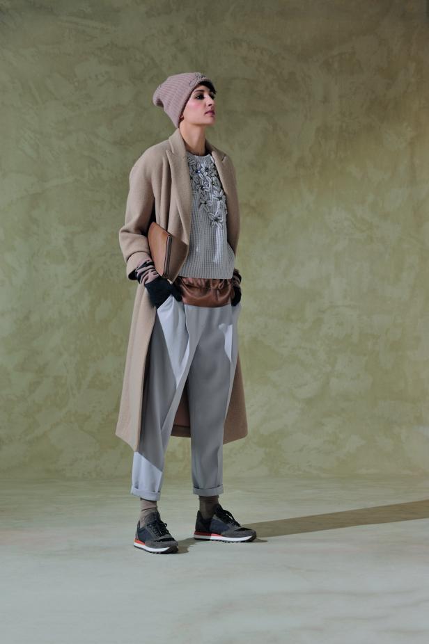 Eine Frau in einem beigen Mantel, einer Mütze und grauen Hosen posiert mit einer Handtasche.