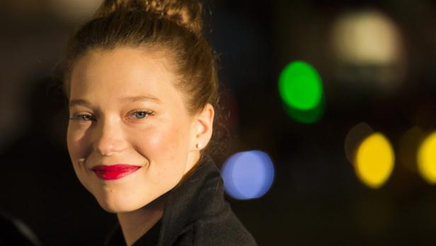 Léa Seydoux lächelt mit rotem Lippenstift vor einem dunklen Hintergrund mit Bokeh.