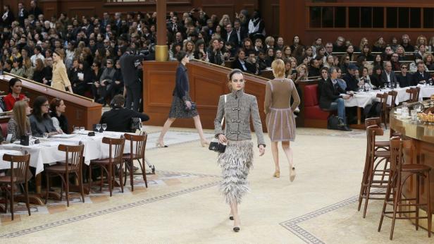 Eine Modenschau mit Models, die vor einem Publikum in einem Café-ähnlichen Ambiente laufen.