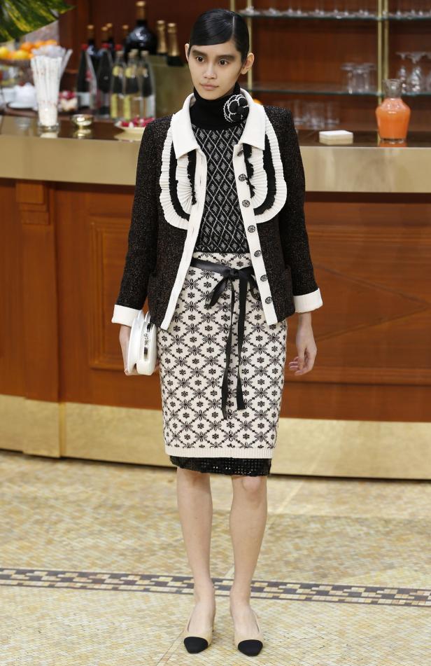 Eine Frau präsentiert ein Outfit mit Tweedjacke und gemustertem Rock vor einer Bar.