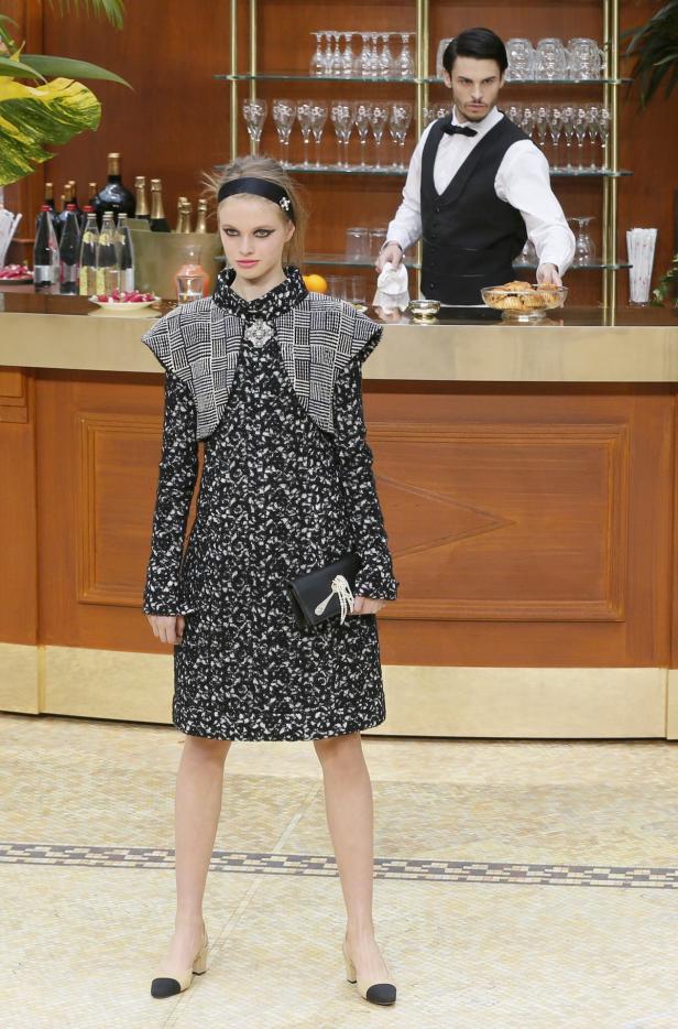 Ein Model in einem schwarz-weiß gemusterten Kleid posiert vor einer Bar, an der ein Barkeeper steht.