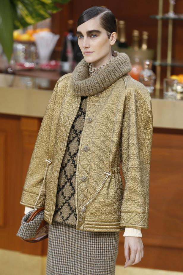 Eine Frau mit streng zurückgekämmten Haaren trägt eine goldene Jacke und einen Rollkragenpullover.