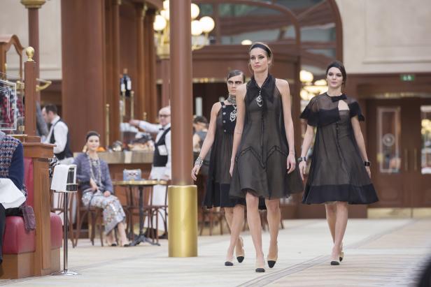 Drei Models präsentieren schwarze Kleider auf einem Laufsteg in einem eleganten Interieur.