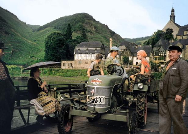 Eine Gruppe von Menschen und ein Traktor auf einer Fähre mit Blick auf ein Dorf und Weinberge.